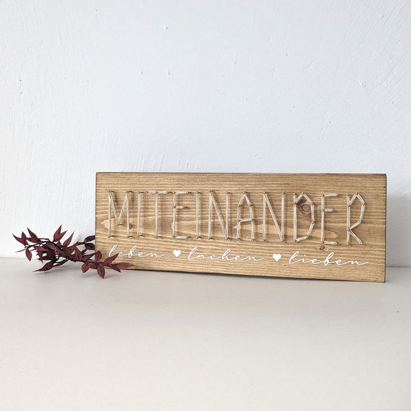 Fadenbild Schriftmotiv „Miteinander“ auf hellem Holzbrett
