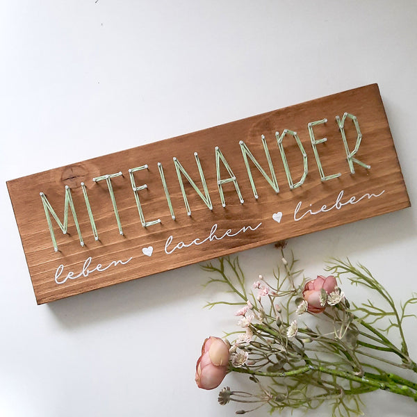 Fadenbild Schriftmotiv „Miteinander“ auf Holzbrett mit Blumen dekoriert