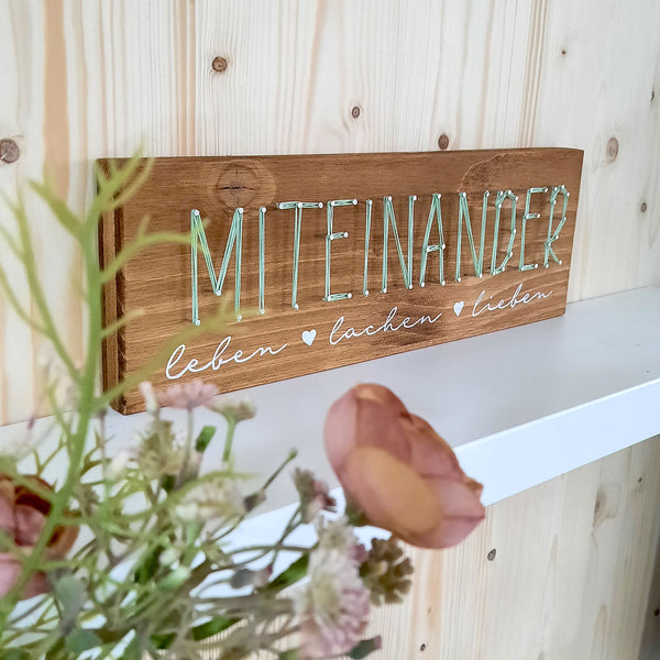 Fadenbild Schriftzug „Miteinander“ als Wanddeko an heller Holzwand
